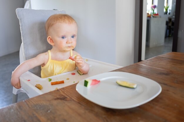 Pensive sweet baby girl trying piece of watermelon while sitting in highchair with food messy on tray and face. First solid food or child care concept