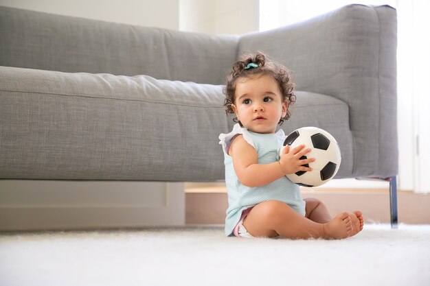 家の床に座って、目をそらし、サッカーボールをしている淡いブルーの服を着た物思いにふける甘い黒髪の女の赤ちゃん。スペースをコピーします。家庭での子供と子供の頃のコンセプト