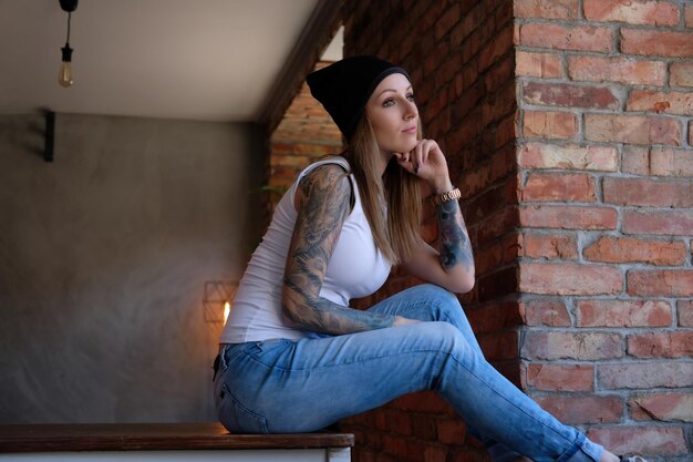 잠겨있는 문신을 한 힙스터 소녀는 흰색 티셔츠와 모자를 쓰고 테이블에 앉아 창 밖을 내다봅니다.
