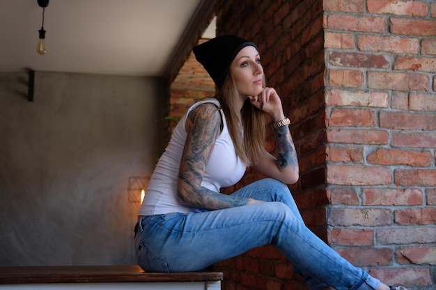 Задумчивая татуированная девушка-хипстер, одетая в белую футболку и шляпу, сидит на столе и смотрит в окно.