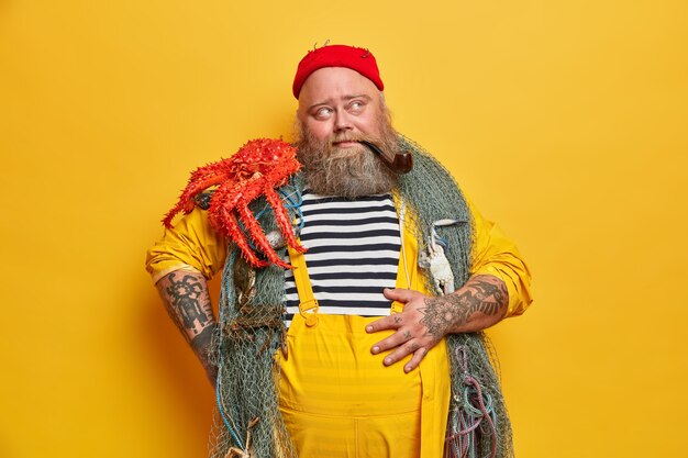 Pensive tattooed sailor keeps hand on belly, carries fishing net over neck, octopus on shoulder, satisfied with successful fishing, takes break, smokes pipe