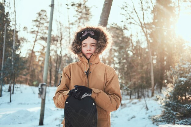 Pensive teen in hood is posing for photographer with his snowboard at winter forest.