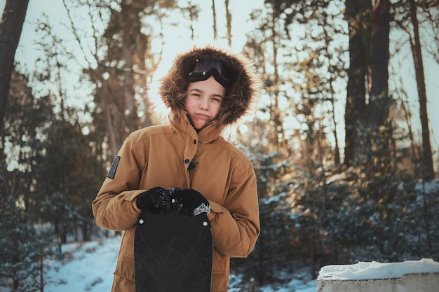 Pensive teen in hood is posing for photographer with his snowboard at winter forest.