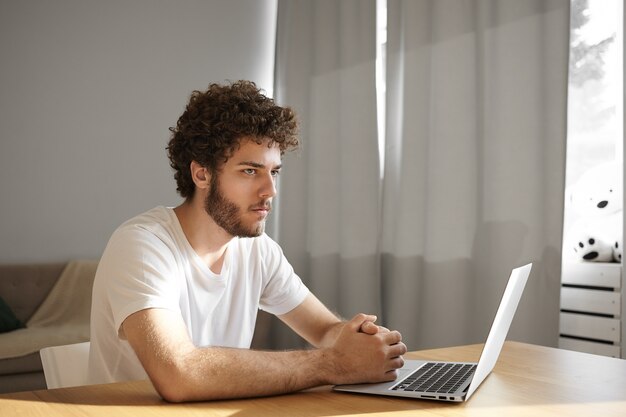 Pensive thoughtful young unshaven male designer with stubble sitting in front of open generic laptop with hands clasped, deep in thoughts while working remotely from home. People and technology