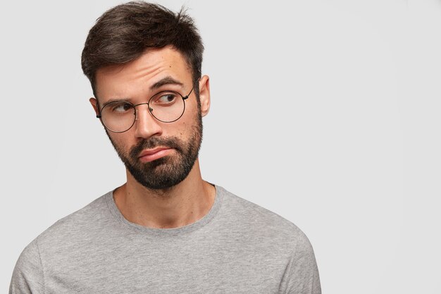 Pensive unshaven attractive male looks thoughtfully aside, has deep thoughts in mind, has round eyewear, poses against white wall