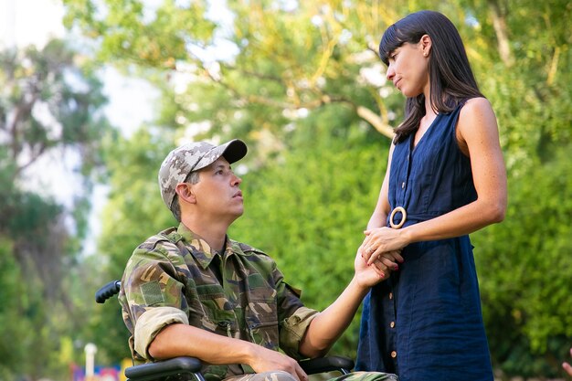 Pensive woman and disabled military man in wheelchair meeting and talking in park outdoors. Disabled veteran or relationship concept