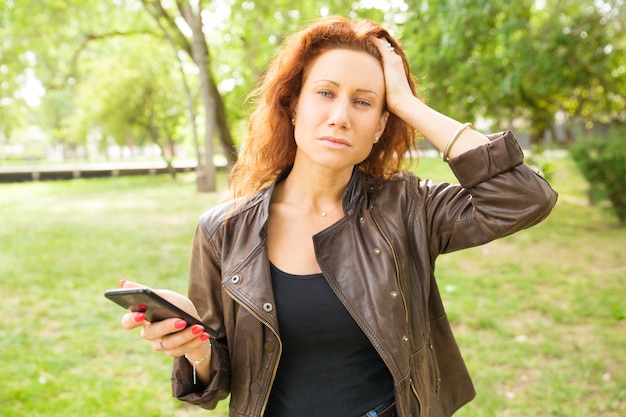 Мечтательная женщина держит мобильный телефон, касаясь головы