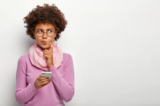 Pensive woman looks thoughtfully aside, holds mobile phone, waits for call, purses lips