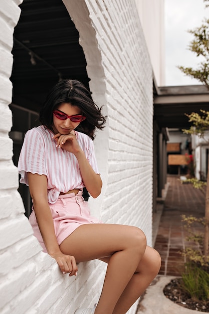 Free photo pensive woman in pink shorts looking down. outdoor shot of tanned female model sitting on bricked wall.