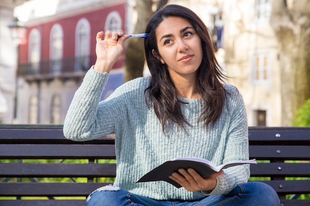 Pensive woman scratching head with pen and sitting on bench