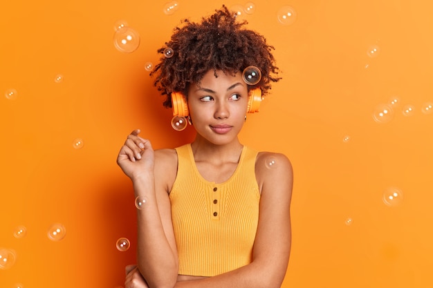 Pensive young afro american woman concentrated aside listens music via stereo headphones has thoughtful expression dressed in casual clothes enjoys lyrics song isolated over orange wall