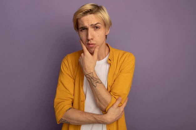 pensive young blonde guy wearing yellow t-shirt putting hand on chin isolated on purple with copy space