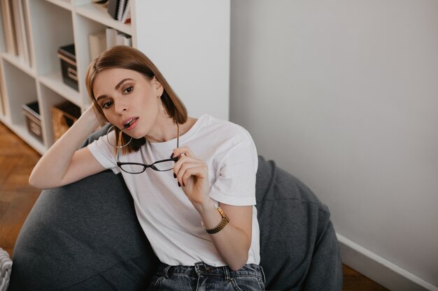 Pensive young girl in white T-shirt took off her glasses. Woman with brown eyes posing sitting in easy chair.