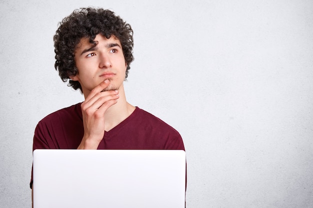 Free photo pensive young male with curly hair, holds chin and looks thoughtfully aside