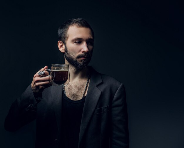 Pensive young man is holding transparent cup of beverage at dark photo studio.