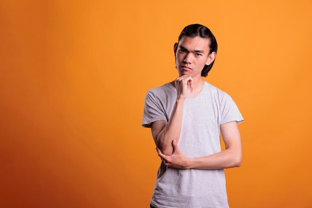 Pensive young man thinking, touching chin with thoughtful facial expression, idea generation. Asian teenager making decision, doubtful person portrait, studio medium shot on orange background