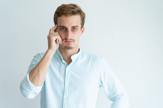 Pensive young man touching head with finger and looking at camera.