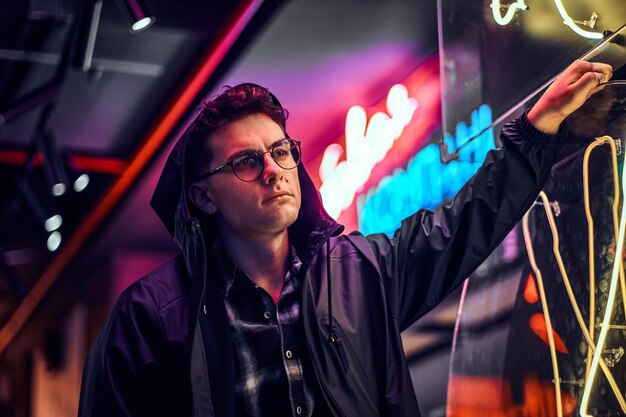 A pensive young man wearing sports hoodie leaning on an illuminated signboard in the street at night.