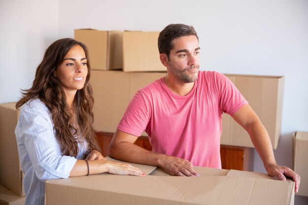 Pensive young man and woman standing among carton boxes in apartment, looking away