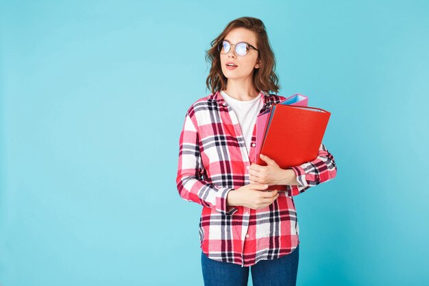 Pensive young pretty lady in eyeglasses and plaid shirt standing with folders in hands and dreamily looking aside on over pink background
