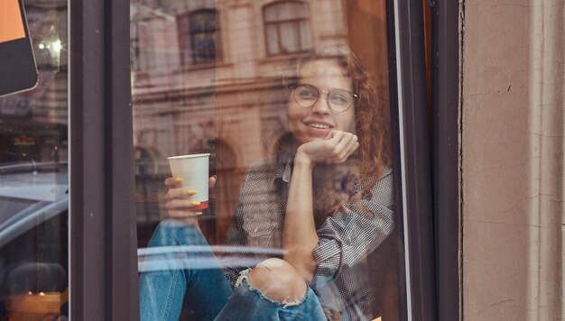 Pensive young redhead curly girl wearing casual clothes and glasses sitting on a window sill with hand on chin, holds a takeaway coffee.