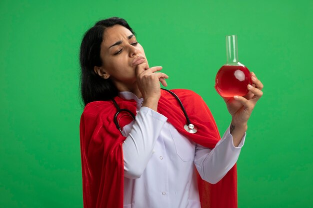 Задумчивая молодая девушка-супергерой в медицинском халате со стетоскопом, держащая и смотрящая на химическую стеклянную бутылку, наполненную красной жидкостью, схватила подбородок, изолированный на зеленом