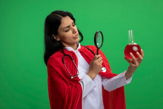 задумчивая молодая девушка супергероя в медицинском халате со стетоскопом, держащая и смотрящая на химическую стеклянную бутылку, наполненную красной жидкостью, с лупой, изолированной на зеленом