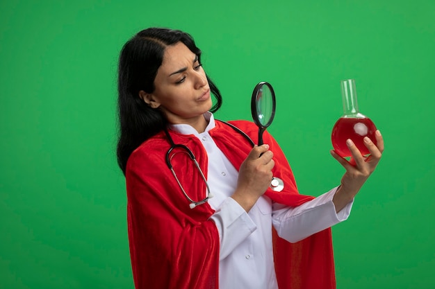 緑に分離された拡大鏡で赤い液体で満たされた化学ガラス瓶を保持し、見ている聴診器で医療ローブを身に着けている物思いにふける若いスーパーヒーローの女の子