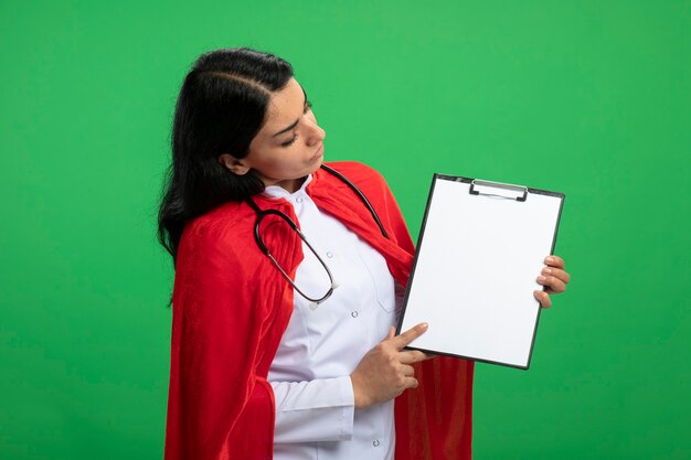 задумчивая молодая девушка супергероя в медицинском халате со стетоскопом, держащая и смотрящая в буфер обмена, изолированную на зеленом