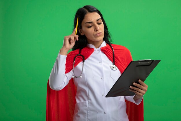 緑に分離された頭にペンを置くクリップボードを保持し、聴診器で医療ローブを着て物思いにふける若いスーパーヒーローの女の子