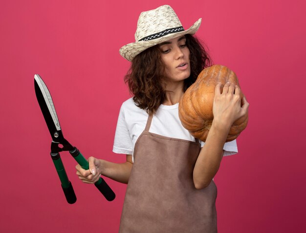 Pensieroso giovane donna giardiniere in uniforme che indossa il cappello da giardinaggio tenendo i clippers e mettendo la zucca sulla spalla isolato sul rosa