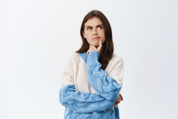 Pensive young woman having doubts, something off, frowning and touching lip while staring thoughtful at upper right corner, standing suspicious against white wall