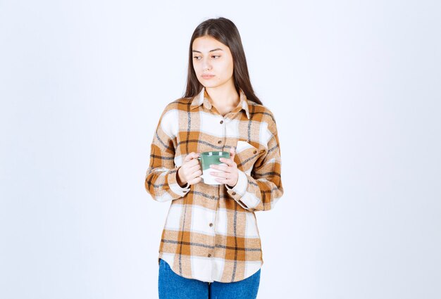 白い壁にコーヒーのカップを保持している物思いにふける若い女性。