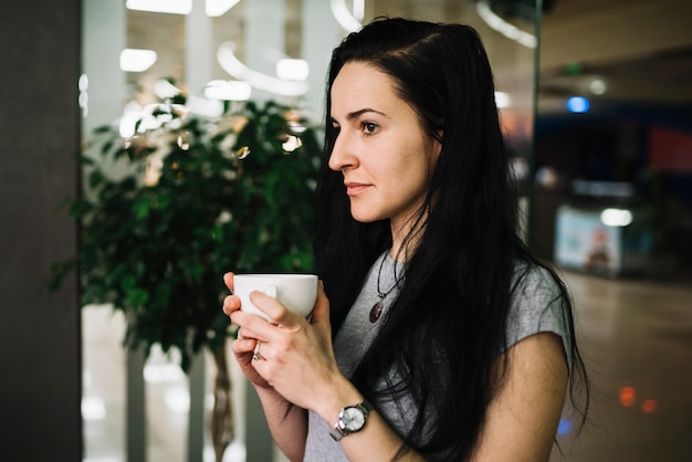 一杯の飲み物を保持している物思いにふける若い女性