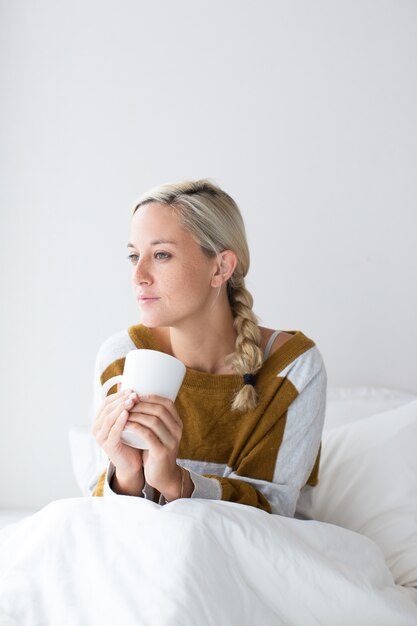 Pensive young woman sitting on bed under blanket