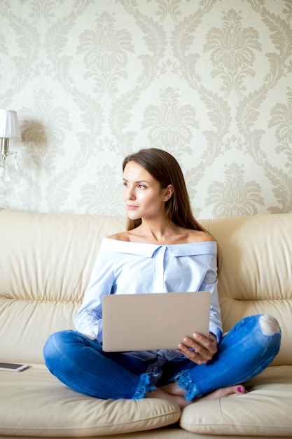 Pensive young woman sitting with laptop on sofa