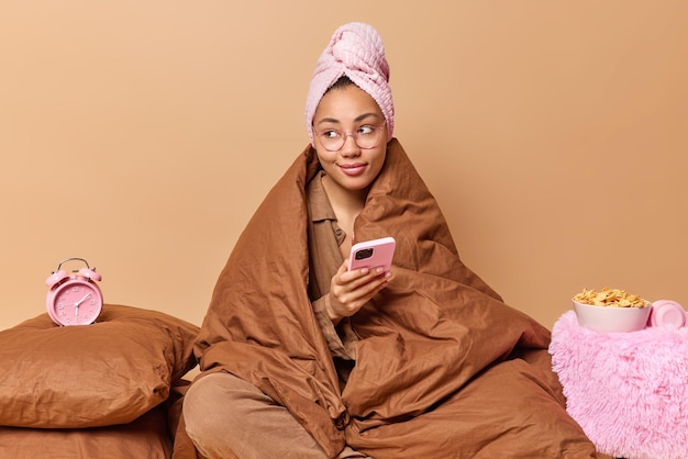 Pensive young woman wrapped in warm blanket has towel on head uses mobile phone sends text messages poses in bedroom near alarmclock wakes up early in morning isolated over beige background