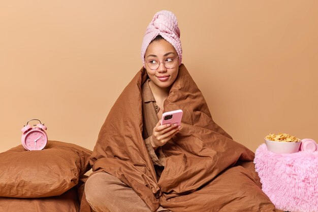 暖かい毛布に包まれた物思いにふける若い女性は頭にタオルを持っています携帯電話はテキストメッセージを送信します目覚まし時計の近くの寝室でポーズはベージュの背景で隔離された朝早く目を覚ます