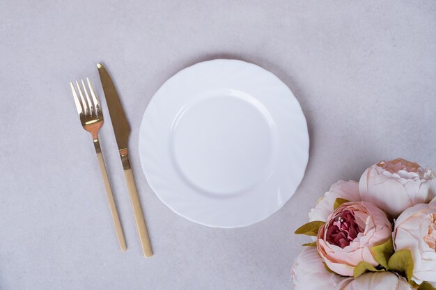 Пионовидные розы, столовые приборы и тарелка на белой поверхности.
