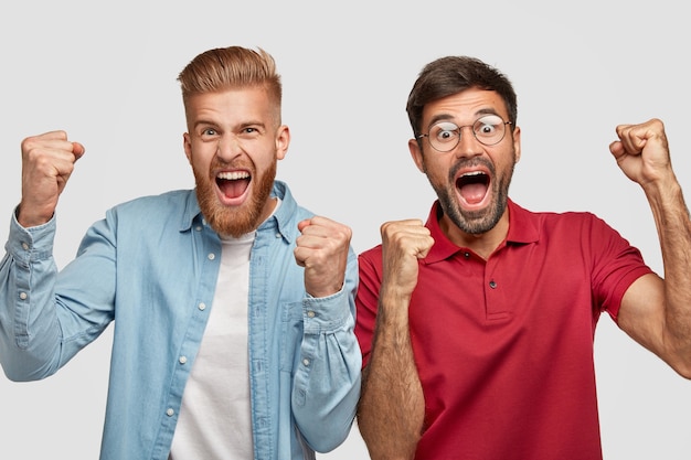 Free photo people, achievement and success concept. best male friends clench fists with happiness, being in high spirit, open mouthes widely, have overjoyed expressions, celebrate their winning, pose indoor