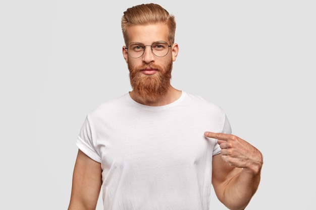 People, advertisement and clothing concept. Serious man hipster with trendy haircut and red beard, indicates at blank space of his t shirt