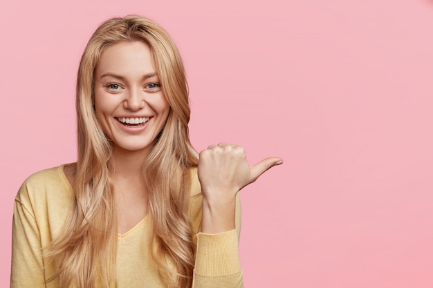 people, advertising and positiveness concept. happy young female with pleasant appearance and smile, indicates with thumb aside at blank pink copy space for your promotional text or hearder.