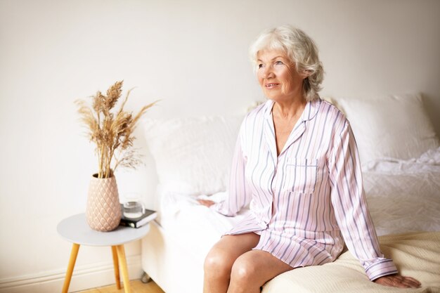 People, age, bedding and bedtime concept. Indoor shot of peaceful relaxed senior retired female sitting on bed in silk pajamas, anticipating beginning of new day. Mature woman going to sleep