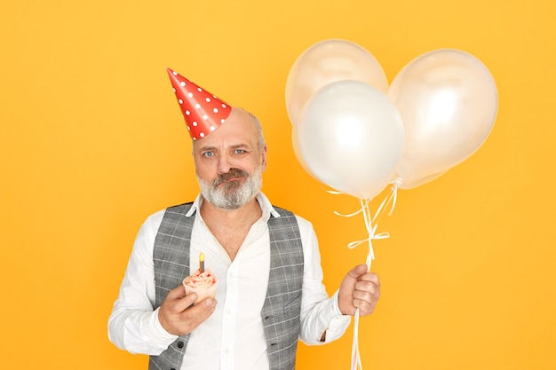 人、年齢、お祝い、休日のコンセプト。気球、コーンハット、カップケーキで隔離されてポーズをとって、彼の引退を祝って、不機嫌そうな表情をしている不機嫌そうな年配のビジネスマンの水平方向のショット