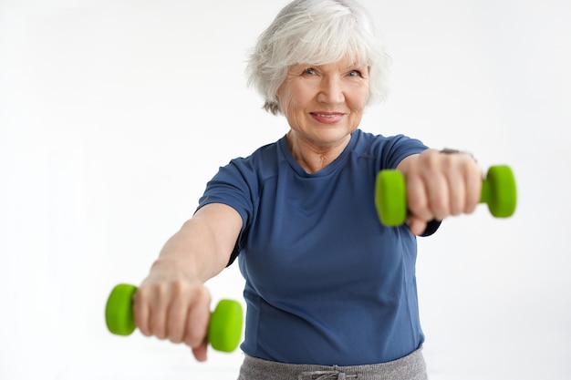 Концепция людей, возраста, энергии, силы и благополучия. Очаровательная улыбающаяся женщина-пенсионерка в футболке делает физические упражнения по утрам, используя пару зеленых гантелей. Выборочный фокус