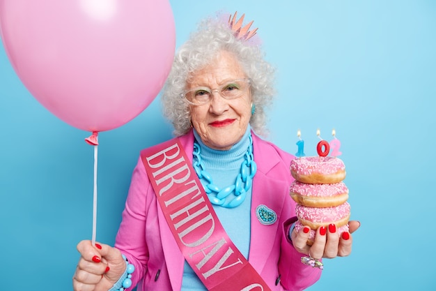 People age holidays festive event concept. Beautiful old woman with curly hair wrinkled face holds glazed doughnuts inflated balloon celebrates bithday