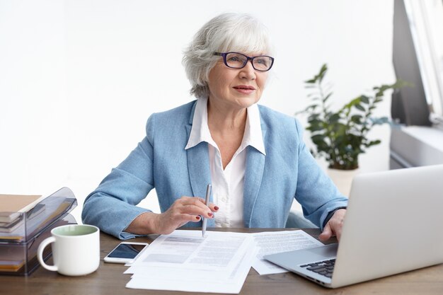 People, age, maturity, job and occupation concept. Indoor shot of beautiful confident elderly female lawyer studying papers and keyboarding on generic portable computer, having thoughtful look