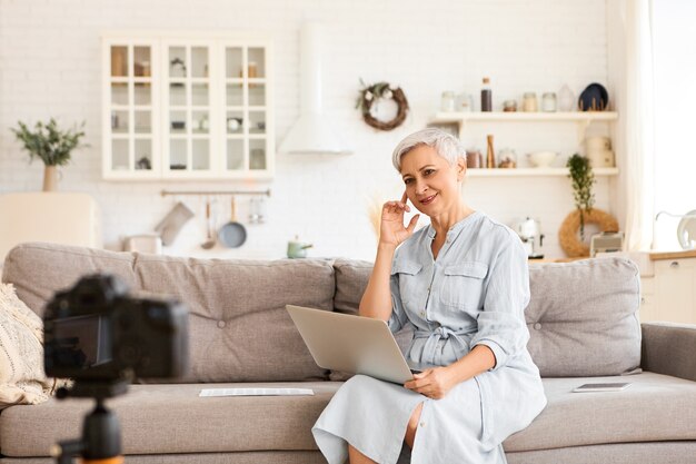 People, age, maturity and modern technology concept. Indoor shot of stylish short haired female blogger recording video on soga in living room, keyboarding on laptop, looking  fixed on tripod