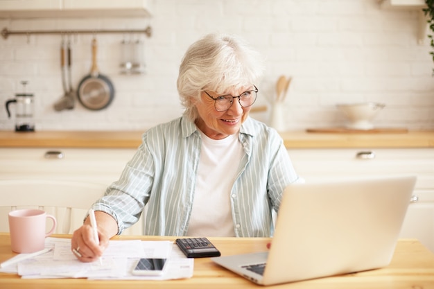 People, age, technology and occupation concept. Indoor image of attractive smiling gray haired woman pensioner using laptop for remote work, sitting in kitchen with papers, earning money online