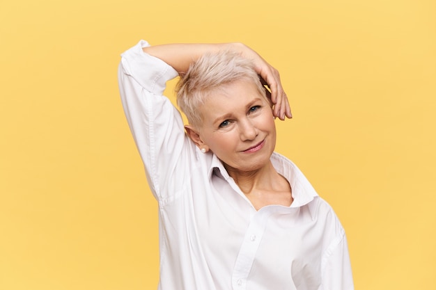 People, aging, maturity, beauty, skin care and health concept. beautiful stylish mature female with pixie dyed haircut bending head and holding hand on cheek doing exercises, smiling